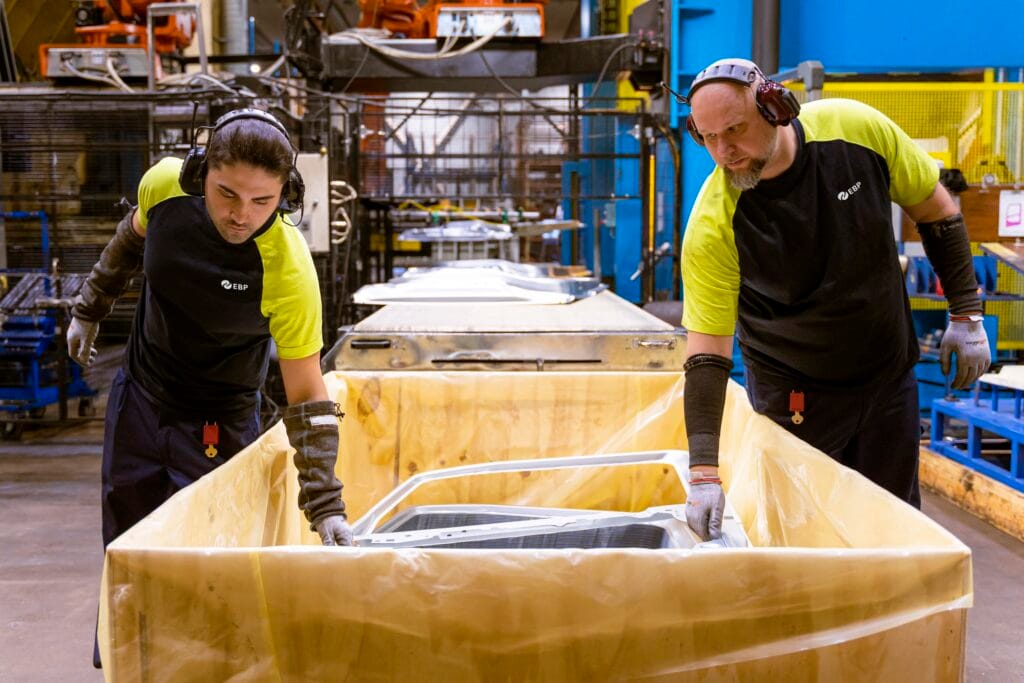 Två operatörer placerar en nypressad karossplåt i en plastfodrad låda vid produktionslinan – plåtpressning i fabriken.